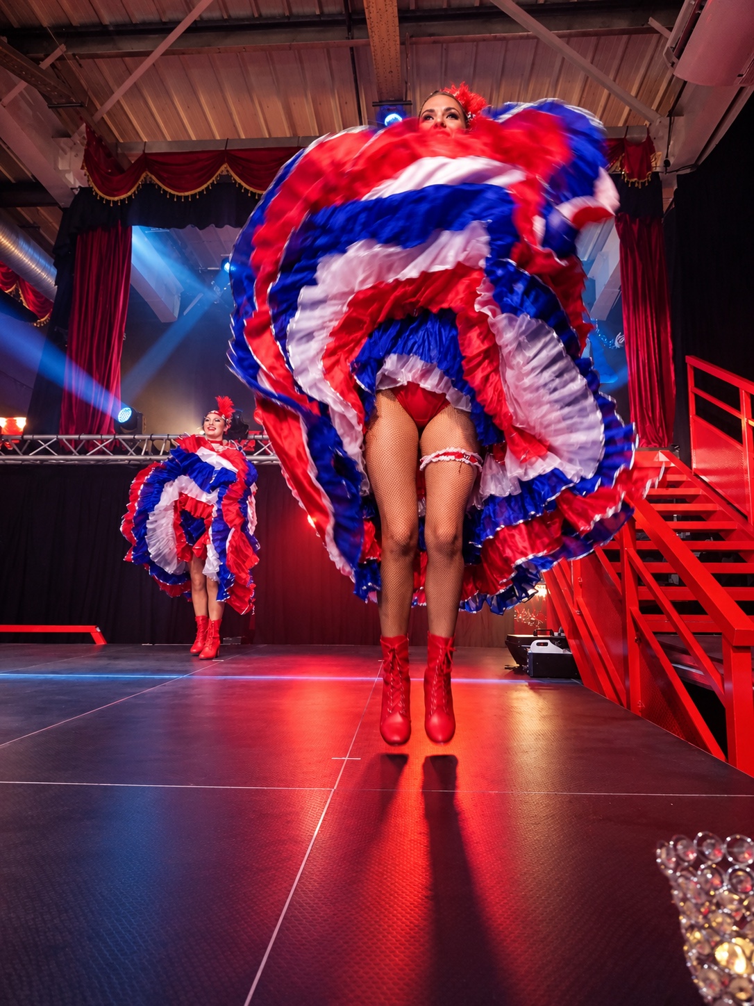 Danseuses de french cancan du Cabaret Dory Arena en costumes bleu, blanc et rouge, avec une artiste au premier plan en plein saut sur scène.